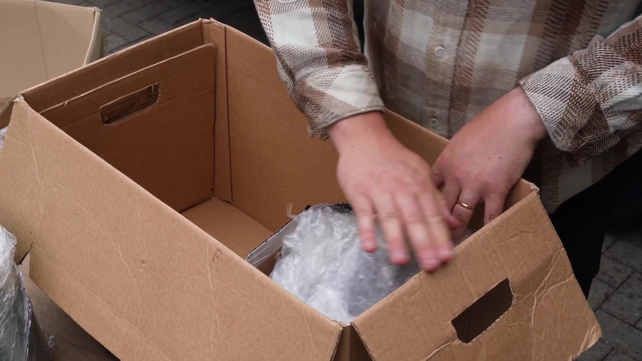 Small business owner arranges products and reused plastic padding inside a cardboard shipping box during order packing.