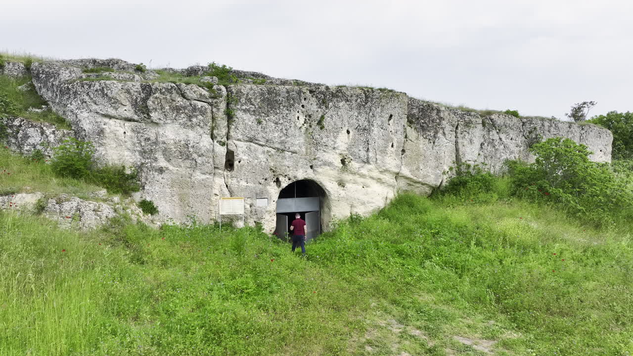Drone orbit of a man walking through tall spring grass toward the arched entrance of a limestone rock-cut medieval church on a green plateau under an overcast sky in Bulgaria