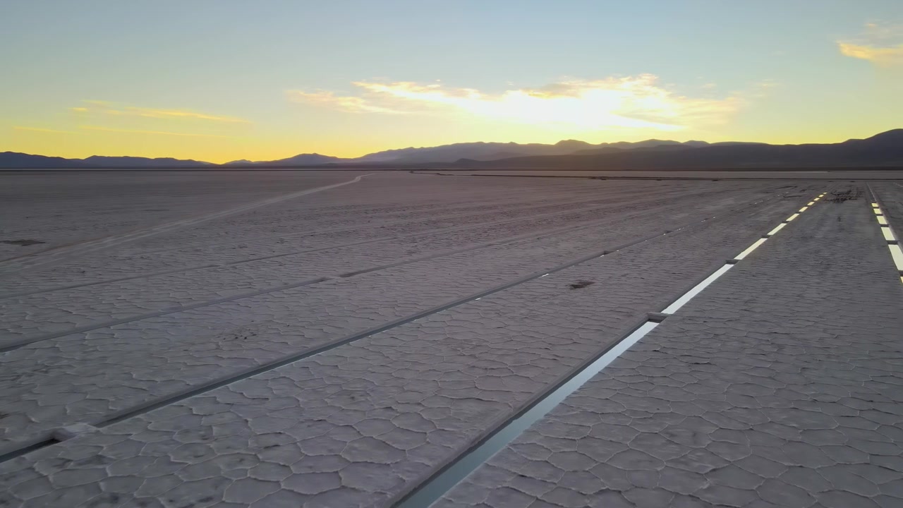 Long narrow channels cut across the cracked surface of Salinas Grandes salt flat as the sun sets behind distant mountains, with a quiet, open desert mood for landscape or travel scenes.