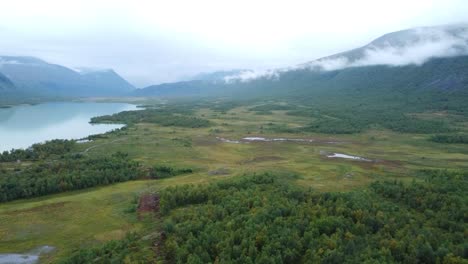 aerial dronw footage flying over sweden green beautiful landscapes with mountains and a large reflective blue lake during a cloudy day while white clouds are between the mountains and forest visible.