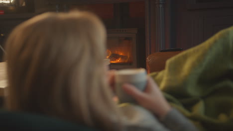 woman at home in lounge lying on sofa looking at cosy fire with hot drink