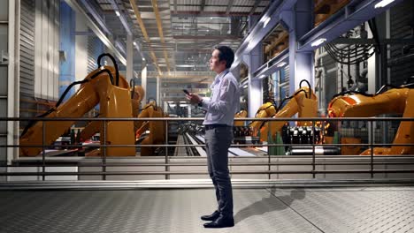 full body side view of asian male worker standing with his phone with industrial robotic arms working, he observes by looking around before he come to concentrating with his smartphone