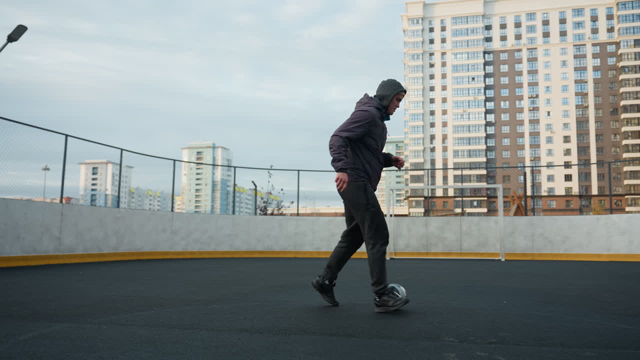 Young athlete skillfully controlling soccer ball with precise footwork during training session on sport arena, background includes urban buildings, fencing, and goalpost under soft daylight