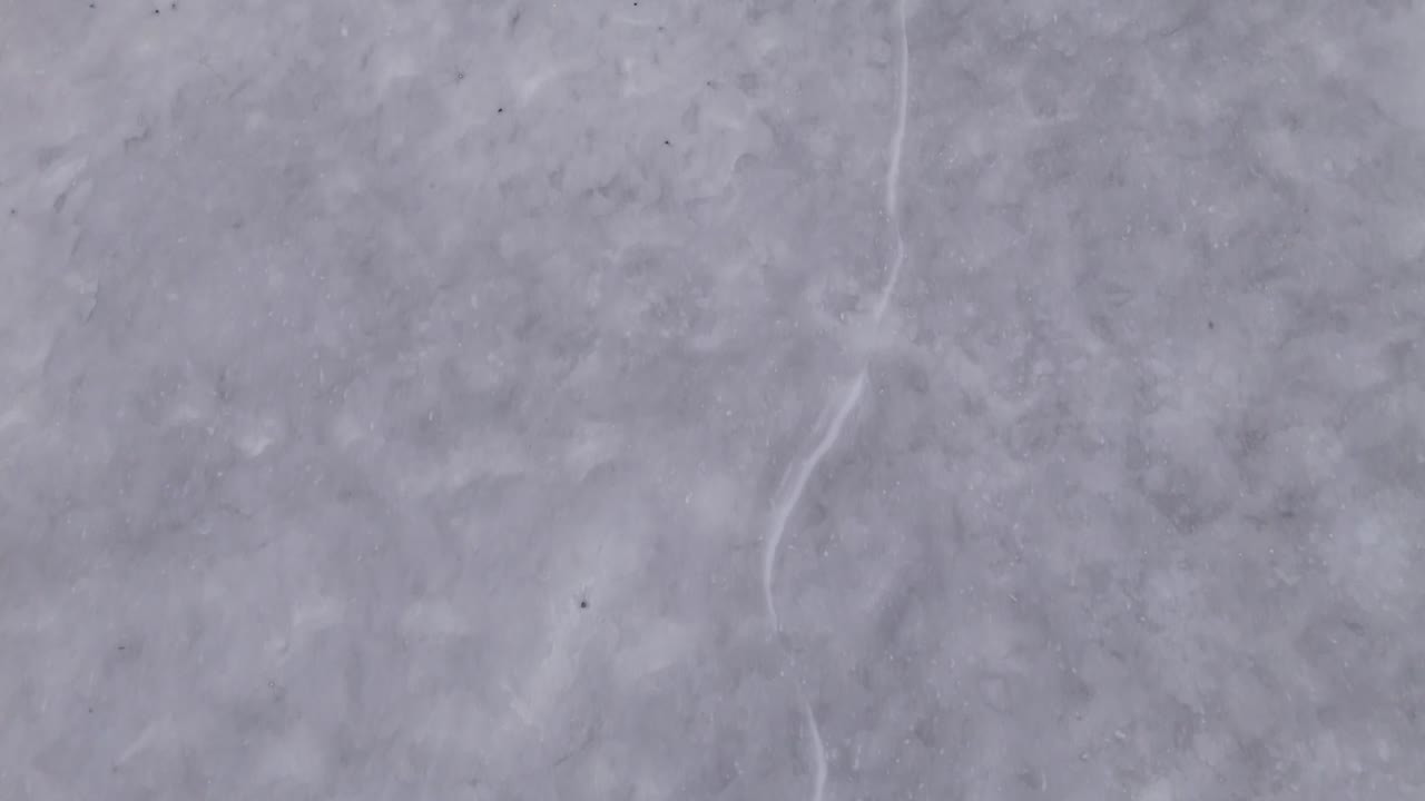 A closeup view of a snowy surface featuring a distinct ice crack. The texture of the snow is clearly visible. Static lockedoff camera