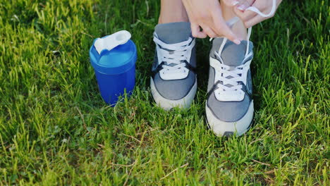 Schnürsenkel-An-Sportschuhen-Weibliche-Beine-Auf-Einem-Grünen-Rasen-In-Der-Nähe-Einer-Flasche-Für-Wasser-Binden,-Die-Für-Eine-Aktivität-Bereit-Ist