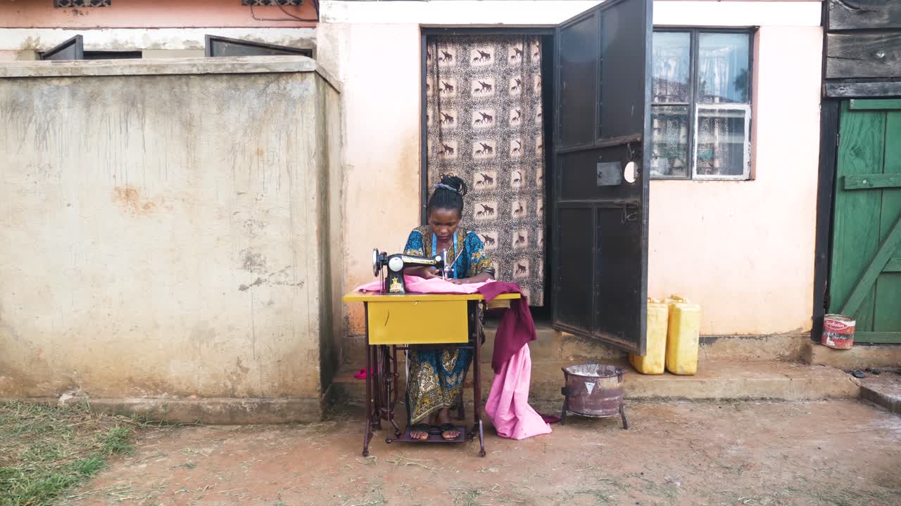 A wide shot in slow motion of an African woman outside her small house sewing clothing on a manual tailoring machine