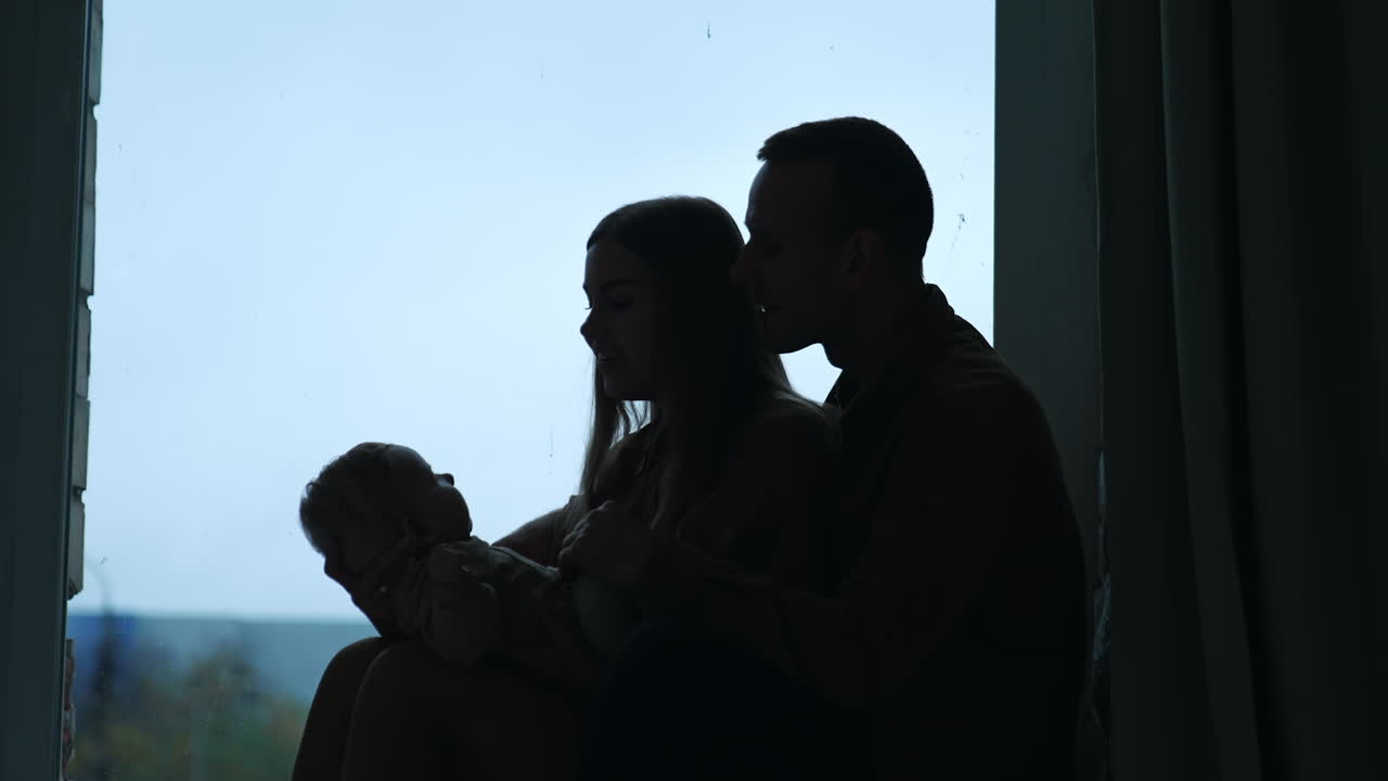Happy parents holding a baby sitting near the window. Silhouettes of loving parents together with their child.