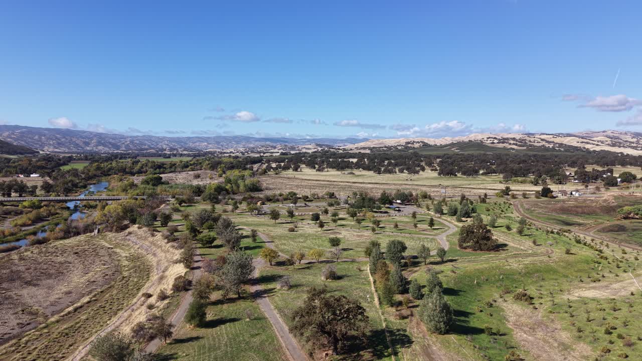 Drone performs a gentle right-to-left pan above winding dirt paths, open fields and Cache Creek, with distant hills under a clear sky in Capay, California