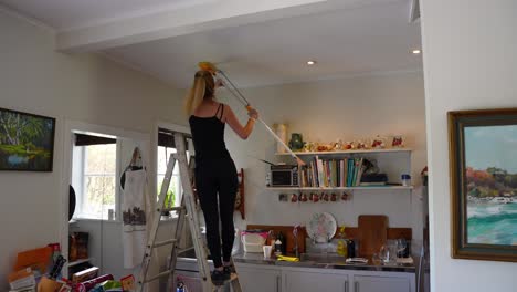 blonde woman stands on the kitchen stairs in a living room cleaning the ceiling