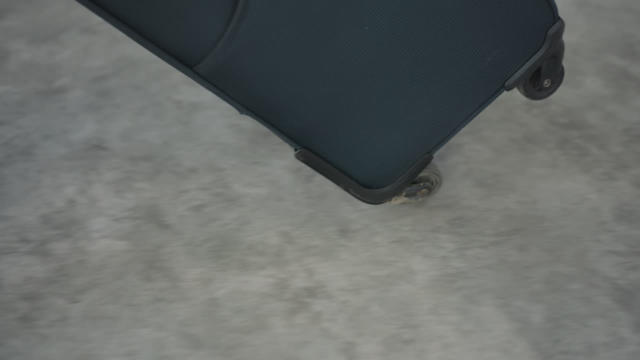Close up low angle view of person dragging wheeled travel bag across smooth floor, pausing to bring bag in front with zipper pocket and handle in focus, casual shoe and pant leg visible