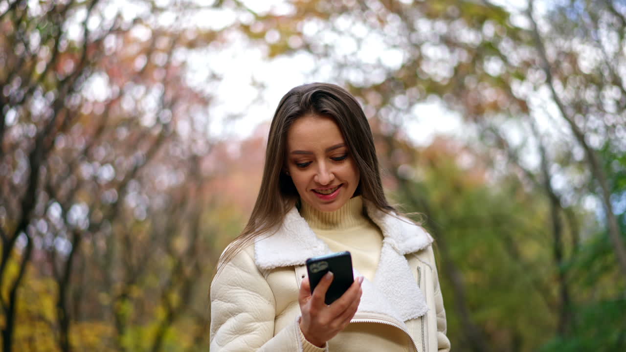 Beautiful smiling Caucasian lady speaks on the phone walking by the park. Lady goes looking at phone and then tries to take a picture of autumn trees.