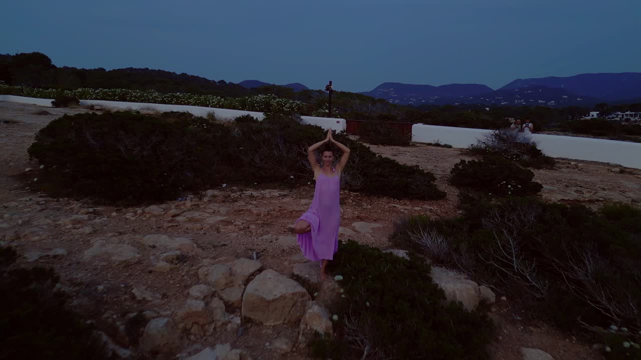 Woman meditating in yoga position on a cliff overlooking the sea at sunset in Ibiza. Nice aerial view flight fly reverse drone
