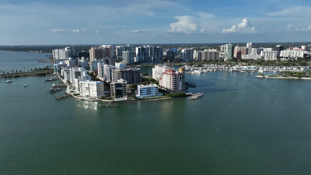 Sarasota, Florida; with a coastal peninsula lined with modern waterfront condos and high-rises, surrounded by calm blue bay waters on a clear, sunny day. Aerial wide shot.