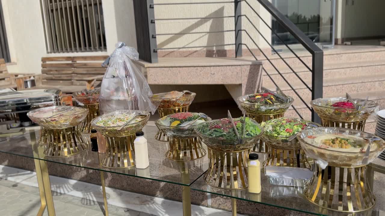 Soleada configuración de catering tipo buffet al aire libre en mesas de cristal con soportes dorados, ensaladeras cubiertas, platos y calentadores junto a las escaleras de un edificio para eventos, bodas o almuerzos de empresa