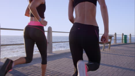 Zwei-Sportliche-Frauen-Laufen-Im-Freien-In-Zeitlupe-Auf-Der-Promenade-Bei-Sonnenuntergang-Am-Meer-Und-Genießen-Den-Abendlauf