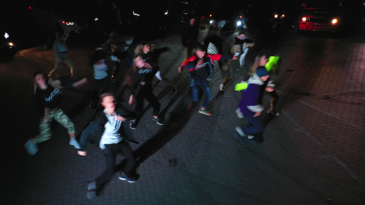 Guys dancing hip hop outdoors at night in the flashlight. Cars with headlights on standing at backdrop. View from above.