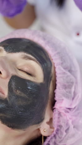a doctor-cosmetologist applies a black mask with a brush to cleanse and moisturize the skin of the face in the beauty center. close-up. vertical video