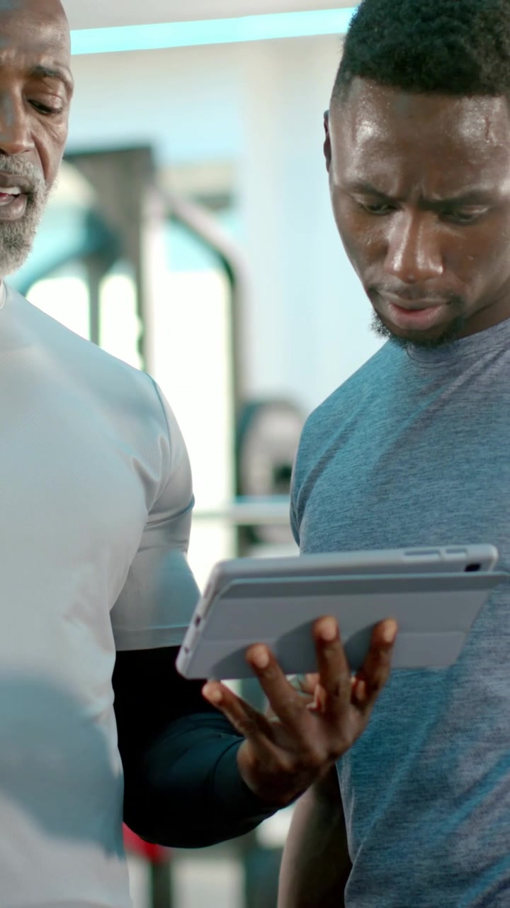 Vertical video: Tapping trainer guiding sweaty trainee wearing tee at gym, reviewing tablet
