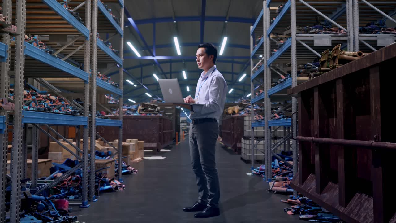 Vista laterale a figura intera di un lavoratore professionista maschio asiatico, in un magazzino industriale di rottami metallici e riciclaggio, che osserva guardando in alto prima di concentrarsi sul laptop e continuare a digitare.