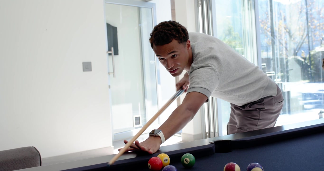 Diverse male friends cheering while lead man leaning over pool table, lining up and striking cue