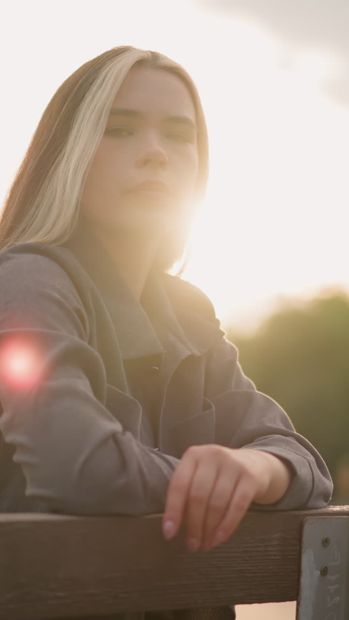 Lady seated at edge of bench, resting head on arm, surrounded by serene outdoor park setting with soft sunlight reflecting around her, background featuring tree and building