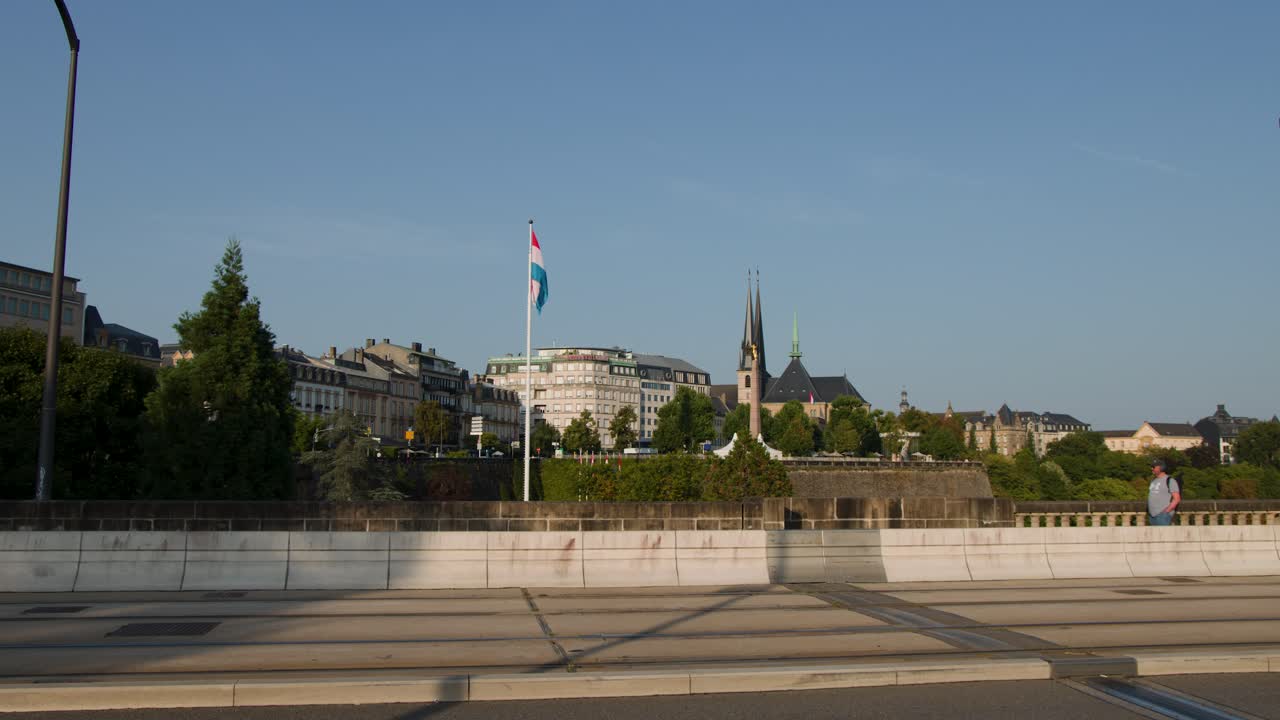 Urban tram and cars traverse city bridge with Luxembourg skyline, cathedral spires, and evening sunlight