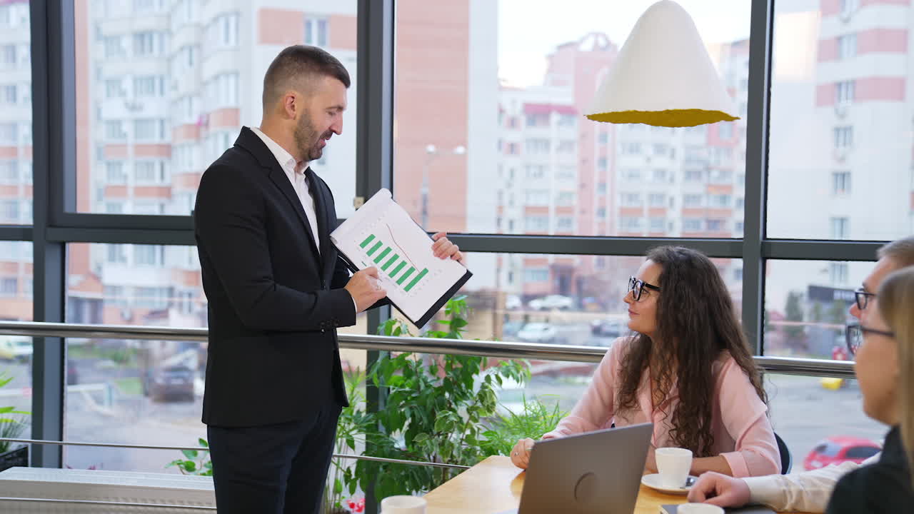 Boss in suit shows and explains the chart to his team. Young employees sit at table, drink coffee and listen to the employer attentively.