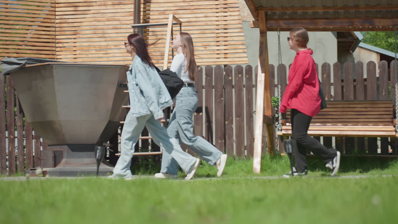 Three young female students walk side by side on grassy path past wooden fence toward warehouse structure, one carrying paper shopping bag, all smiling under bright sunlight