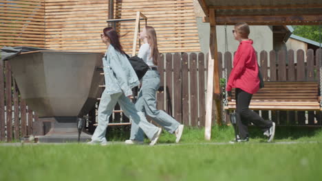 three young female students walk side by side on grassy path past wooden fence toward warehouse structure, one carrying paper shopping bag, all smiling under bright sunlight