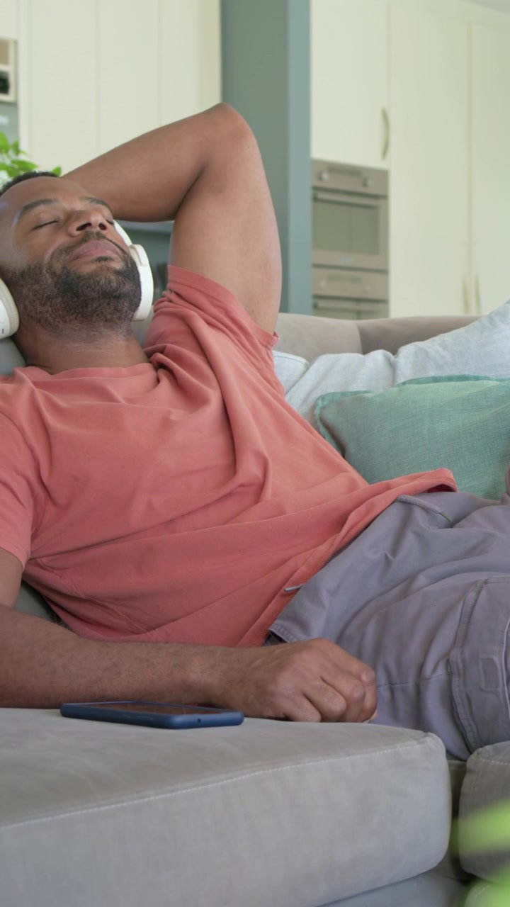 Video verticale: Uomo sorridente reclinato in soggiorno, cuffie che riproducono musica, smartphone sul divano