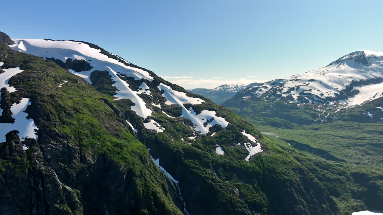 Snow reflecting bright sunlight lies on the mountain slopes. Drone footage over the spectacular rocky landscape of Alaska, USA