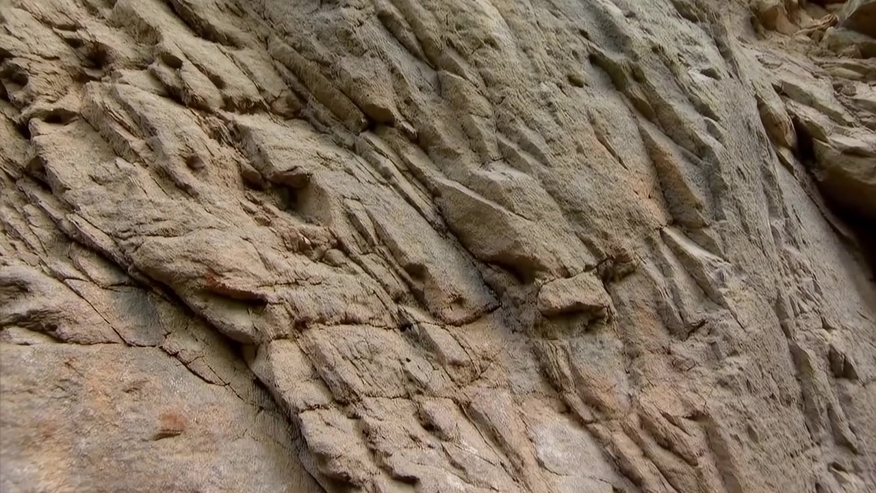 Lavvinklet udsigt over forvitret kystklippevæg, der viser diagonale striber, lagdelt erosion, rygge, furer, lavvandede lommer, ru tekstur og bløde dæmpede jordfarver med en subtil naturlig overflade