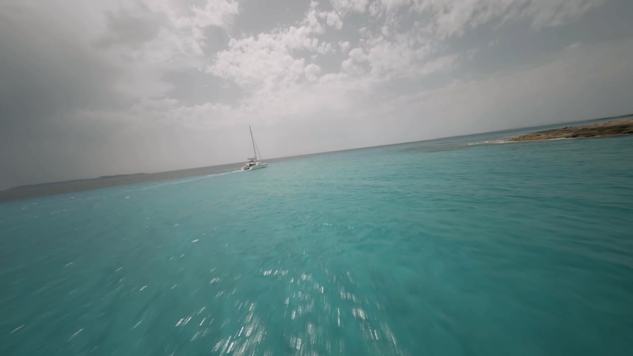 toma de un dron fpv de un velero blanco navegando sobre una hermosa agua de mar de color turquesa a lo largo de una playa rocosa en un día soleado