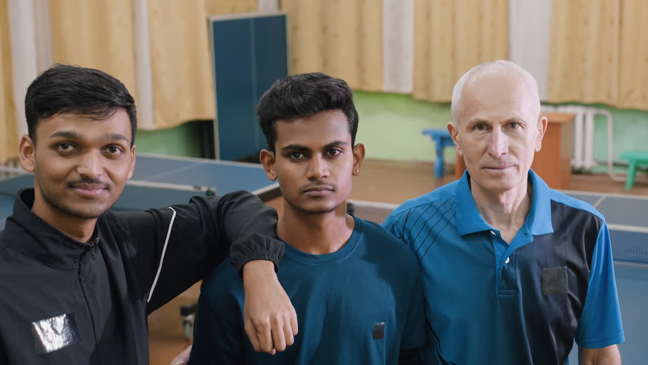 Jugadores de tenis posando con el entrenador en el interior después del partido, con los brazos entrelazados mostrando trabajo en equipo, disciplina, asesoramiento, orgullo, determinación y espíritu deportivo durante la sesión de fotos en el pabellón de entrenamiento.
