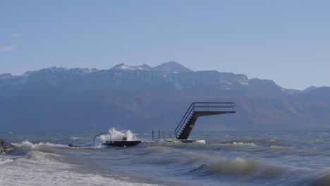 Sprungbrett-An-Einem-Windigen-Tag-Mit-Wellen,-Die-Auf-Die-Struktur-Schlagen,-Die-Schneebedeckten-Alpen-Im-Hintergrund