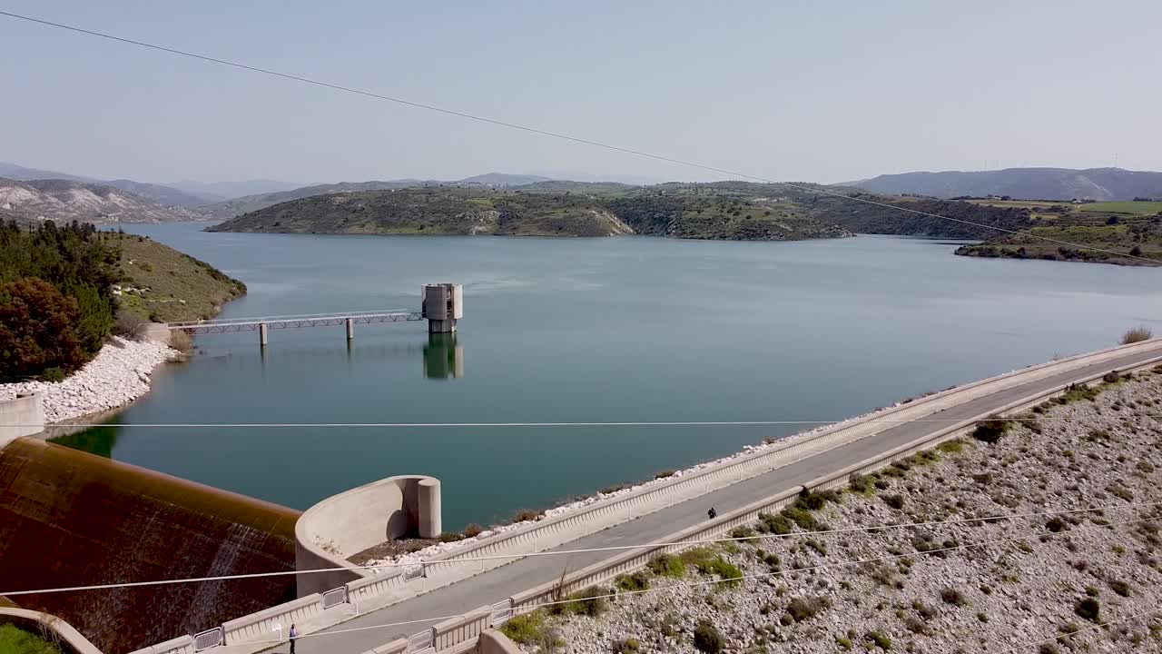 A tranquil view of a large reservoir surrounded by green hills on a sunny afternoon showcases the beauty of nature. Water reflects the clear sky, creating a peaceful atmosphere