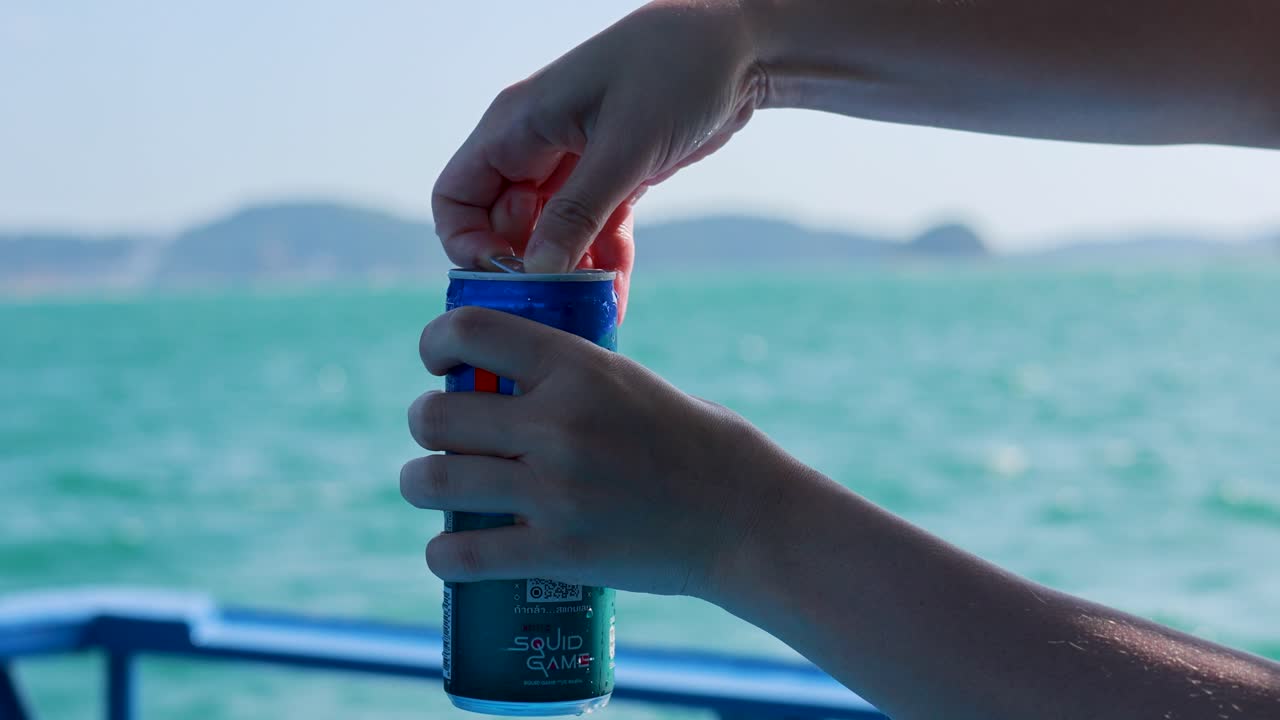 Hands opening a soda can on a boat with turquoise ocean and distant islands in the background, under bright daylight
