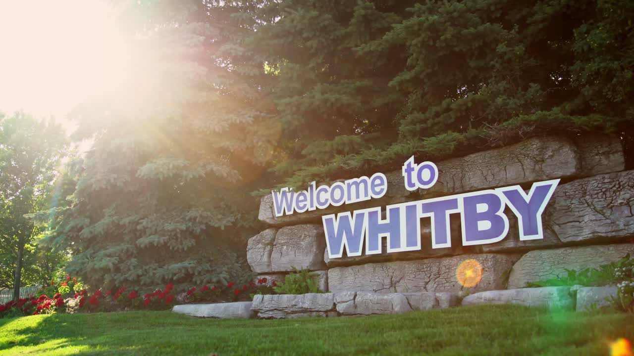 "Welcome to Whitby" is prominently displayed on a sign affixed to a stone wall beneath a canopy of trees, as the camera smoothly pans to capture the scene