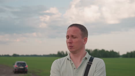 man in light grey shirt with black shoulder strap standing on rural road next to open green field communicating with distant parked car under cloudy sky