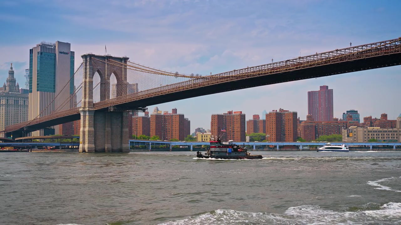 Rimorchiatore che naviga sull'East River sotto il Ponte di Brooklyn a New York. Vista laterale di una barca scura nell'acqua, struttura sospesa in acciaio sopra, edifici in mattoni, illuminazione diurna.