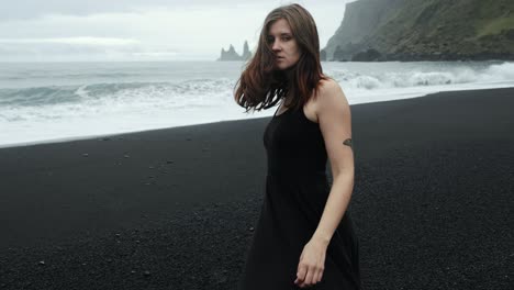 young beautiful woman in black dress dancing on black sand beach iceland, slow motion walking, looking into camera