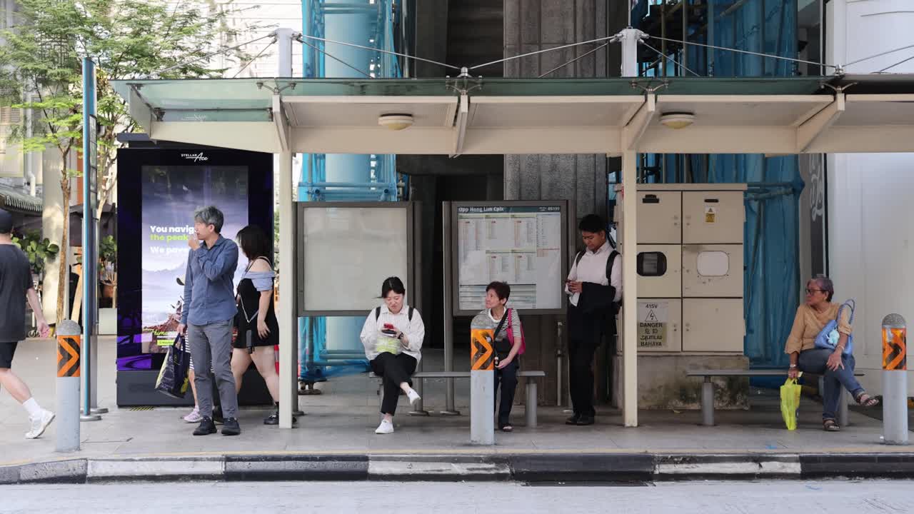 Several individuals wait and interact at a modern Singapore bus stop in daylight, with natural lighting and a static, wide camera angle capturing urban street life