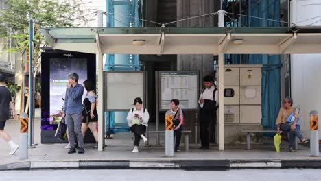 several individuals wait and interact at a modern singapore bus stop in daylight, with natural lighting and a static, wide camera angle capturing urban street life