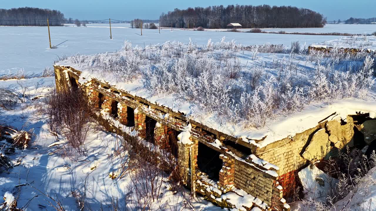 Droonikuvassa vanha, tuhoutunut rakennus, joka on valkoisen kuuran ja lumen peitossa latvialaisella maaseudulla aurinkoisena talvipäivänä. Sininen taivas ja lumiset pellot on valaistu auringonnousun valolla.