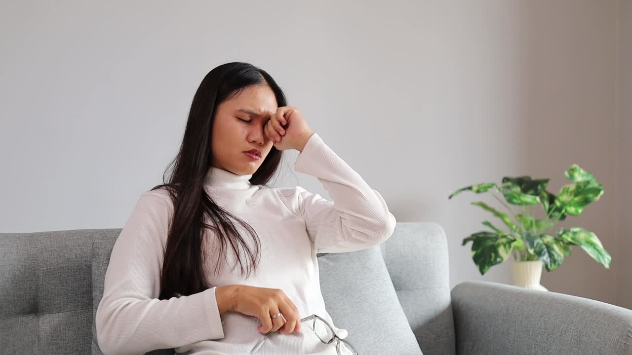 Seorang wanita Asia yang lelah dengan kemeja putih menggosok matanya saat beristirahat di sofa. Sebuah momen realistis dan emosional tentang kelelahan serta kejenuhan digital dalam gaya hidup modern