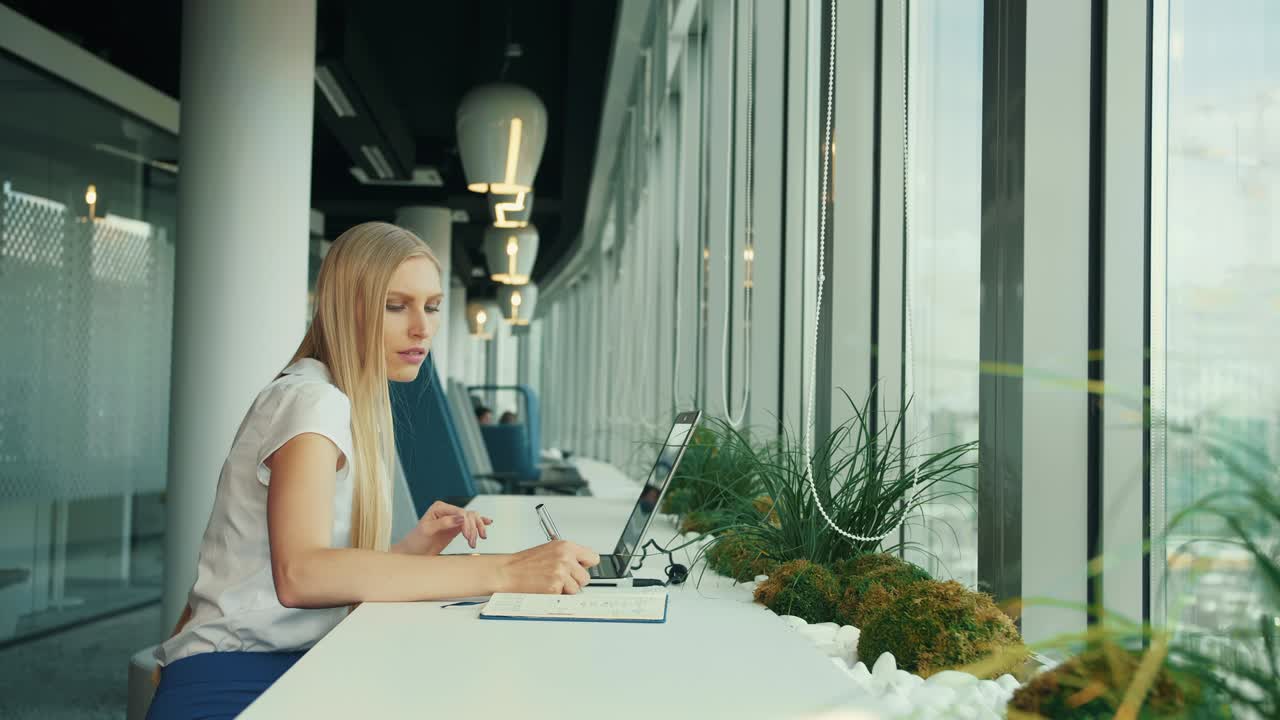 Donna d'affari che lavora con il laptop nel nuovo ufficio. Vista laterale di una donna seduta al tavolo accanto alla finestra in un ufficio moderno e che usa il laptop alla luce del giorno