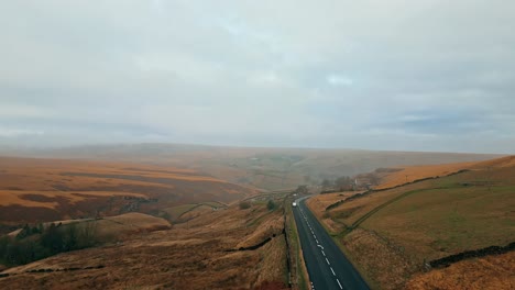 Carretera-Que-Cruza-El-Sombrío-Páramo-Expuesto-Y-Azotado-Por-El-Viento-Sobre-Huddersfield,-Yorkshire,-Inglaterra-Con-Un-Solo-Coche-Que-Viaja-A-Lo-Largo