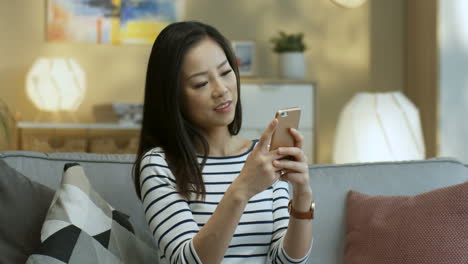 Mujer-Joven-Con-Blusa-A-Rayas-Tocando-Y-Enviando-Un-Mensaje-De-Texto-En-El-Teléfono-Inteligente-Sentado-En-El-Sofá-En-La-Sala-De-Estar