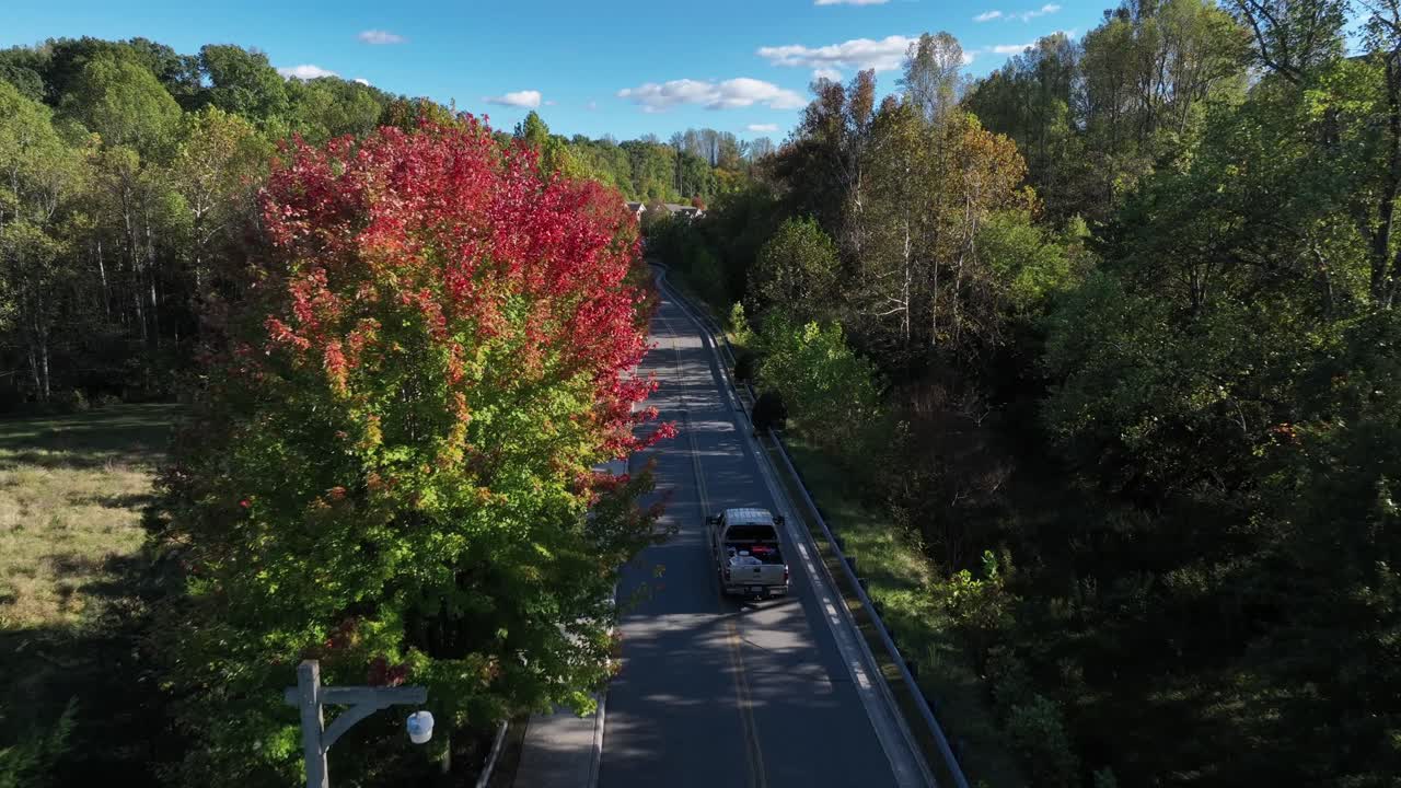 Aerial tracking shot of Pickup Truck on rural road between multi-colored trees. Beautiful sunny day in fall season. American interstate road in suburbia of Pennsylvania, USA
