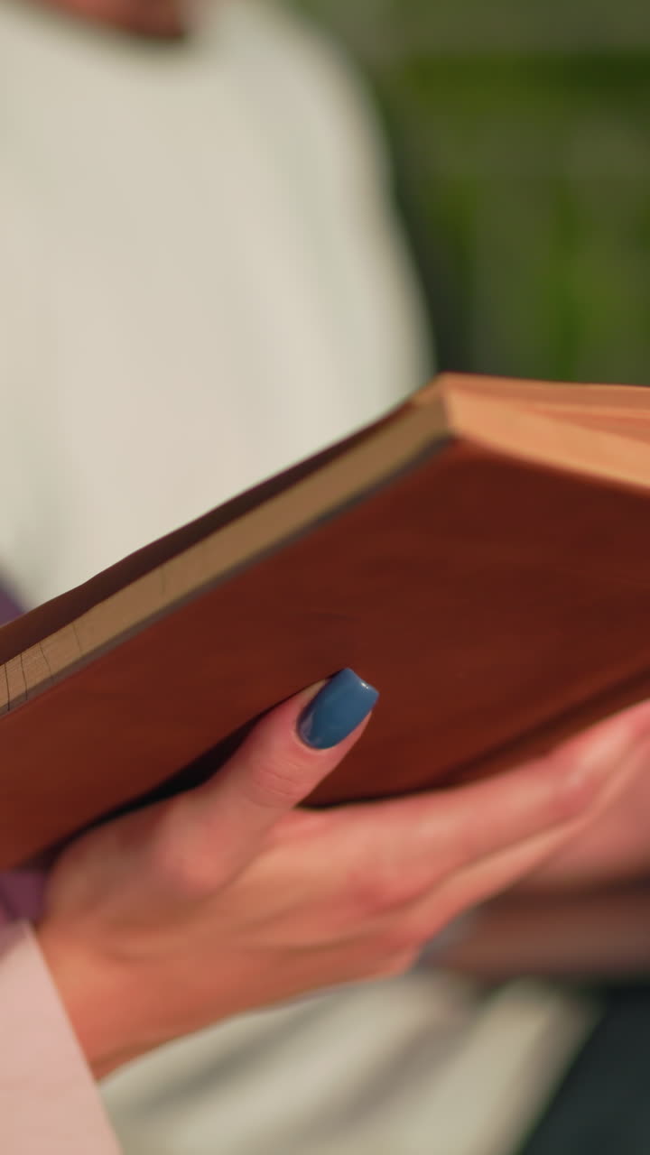 primer plano de una mujer en rosa leyendo un libro con uñas pintadas de azul mientras su amiga en el fondo escribe en una computadora portátil, ligeramente borrosa, la escena incluye una colina verde, un poste y un entorno al aire libre relajado